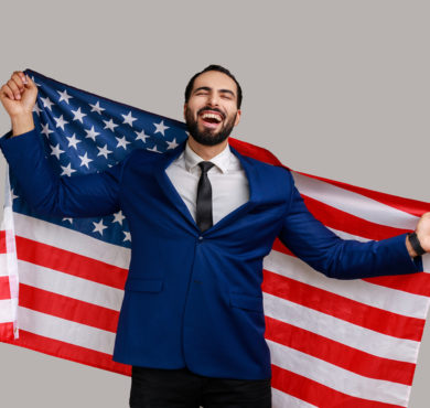 Excited Bearded Man Holding Usa Flag And Looking At Camera With Excited bearded man holding USA flag and looking at camera with rejoice look, celebrating national holiday, wearing official style suit. Indoor studio shot isolated on gray background.